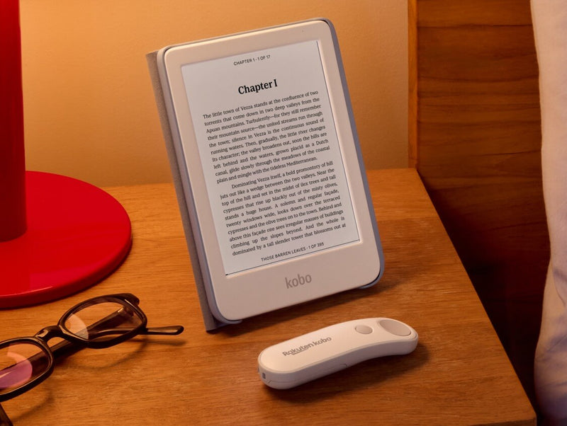 A white Kobo Clara Colour is propped up on table in its SleepCover with built-in stand, alongside a white Kobo Remote and a pair of eyeglasses.